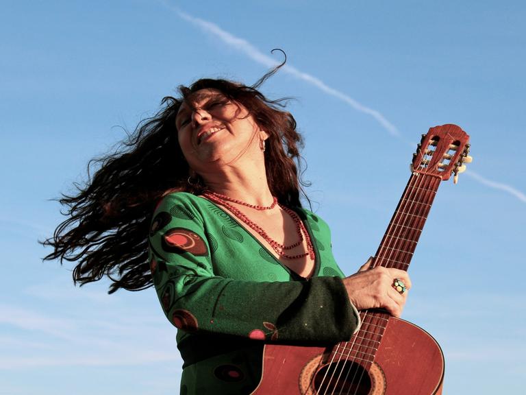 Zélia Fonseca dreht sich schwungvoll mit ihrer Gitarre und offenem Haar vor blauem Himmel. 