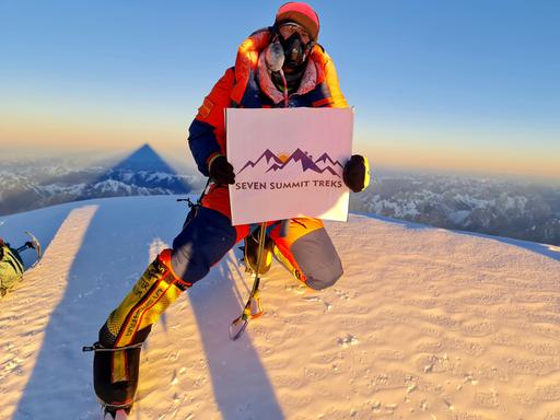 Ein Bergsteiger auf einem schneebedeckten Gipfel hält ein Plakat mit der Aufschrift "Seven Summit Treks".