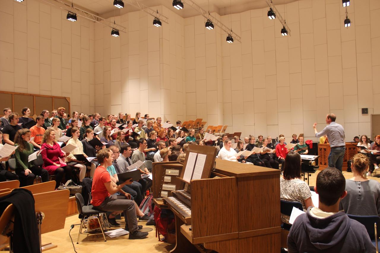 In einem nüchtern erleuchteten Saal singt ein Chor bestehend aus jugendlichen Sängerinnen und Sänger angeführt von einem Dirigenten. In einem nüchtern erleuchteten Saal singt ein Chor bestehend aus jugendlichen Sängerinnen und Sänger angeführt von einem Dirigenten.