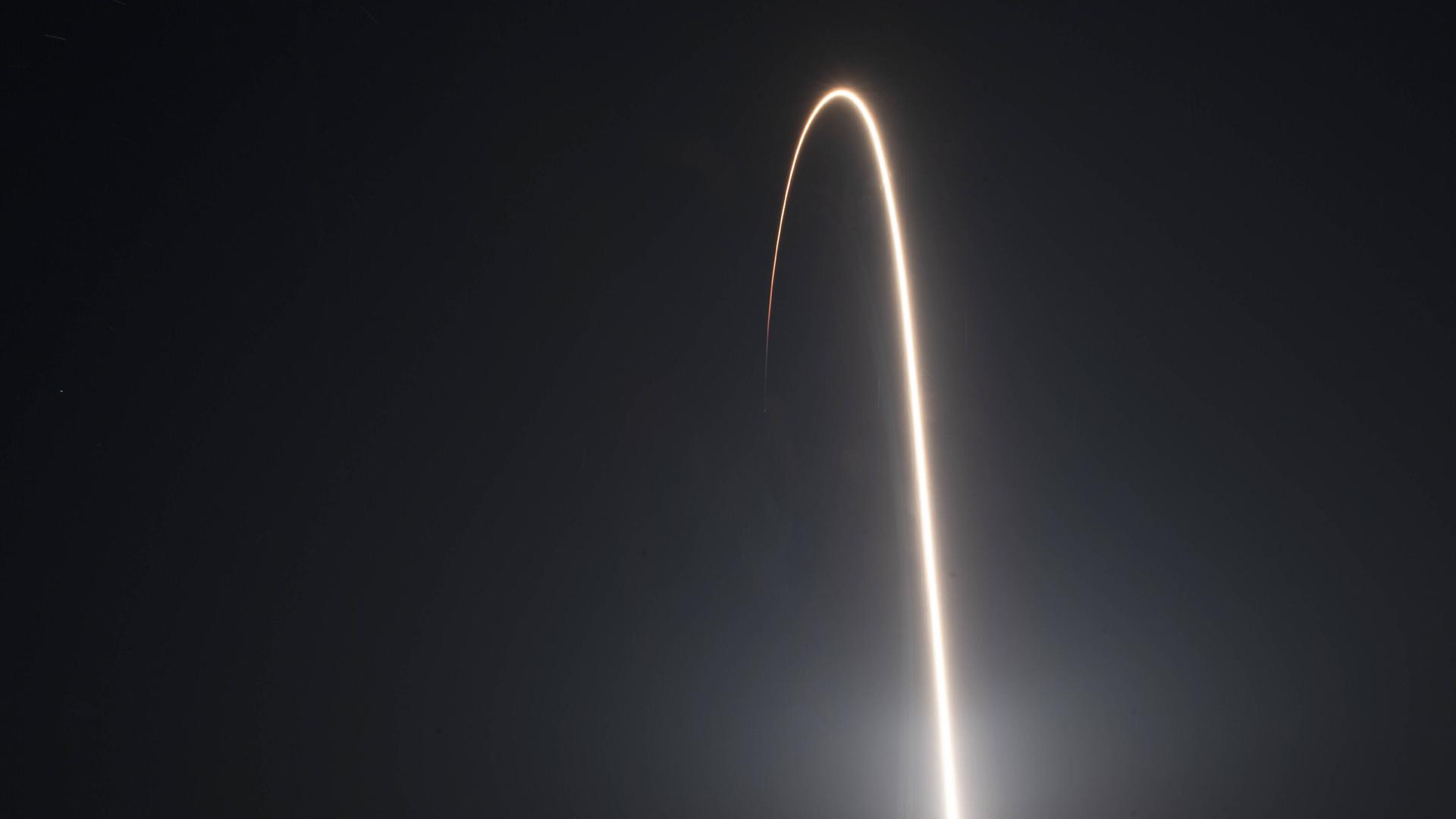 Langzeitbelichtung eines Raketenstarts, bei dem die Rakete einen langen, gebogenen Schweif in der Form der Flugbahn hinterlässt.