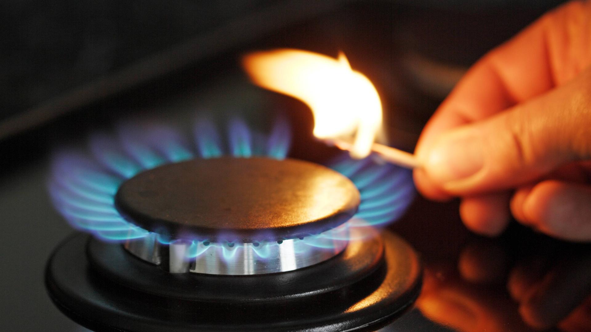 Eine Hand hält ein brennendes Streichholz an eine Flamme am Gasherd.