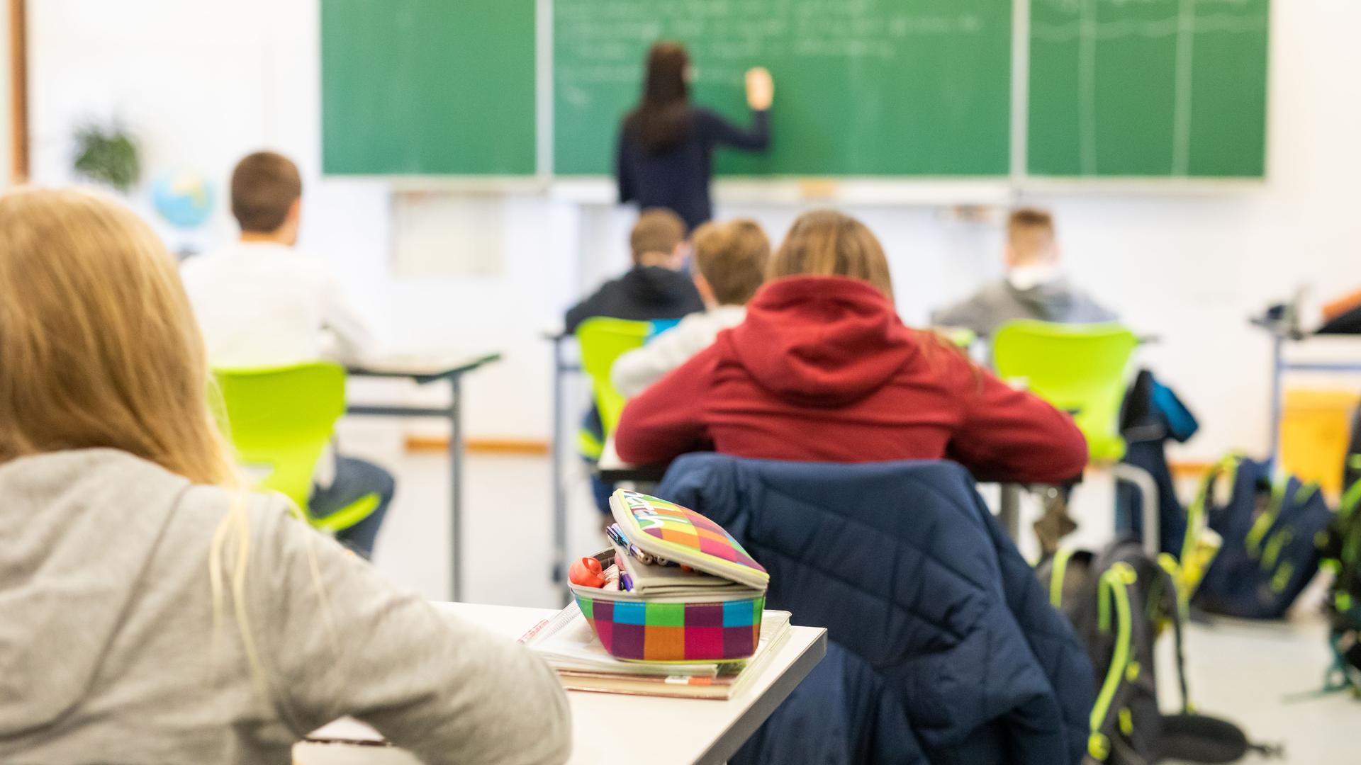 Schülerinnen und Schüler sitzen im Unterrichts in ihrem Klassen-Zimmer. Eine Lehrerin steht an der Tafel.