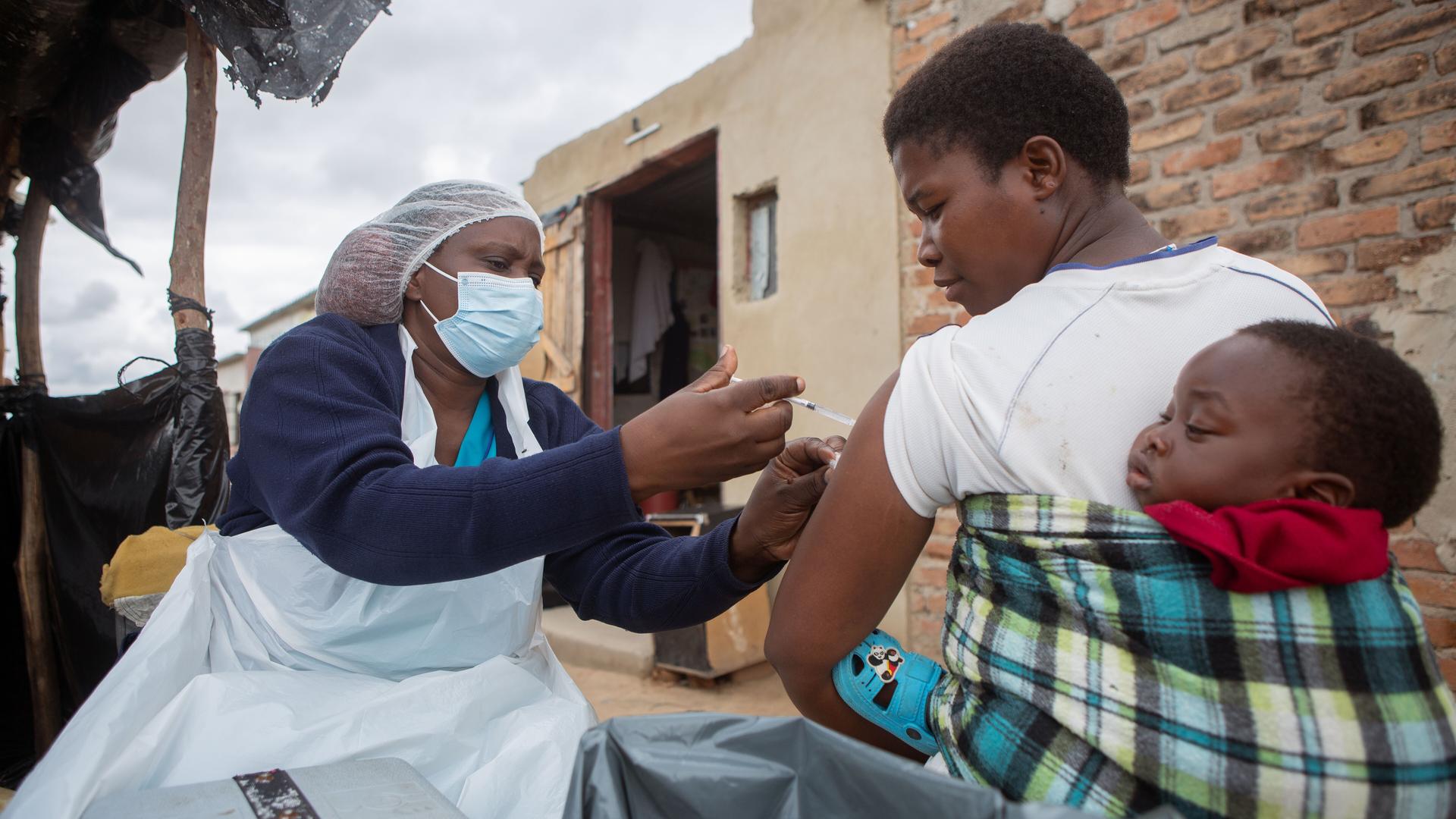 Eine Krankenschwester impft in Zimbabwe eine Mutter, die ihr Kind auf dem Rücken trägt.  