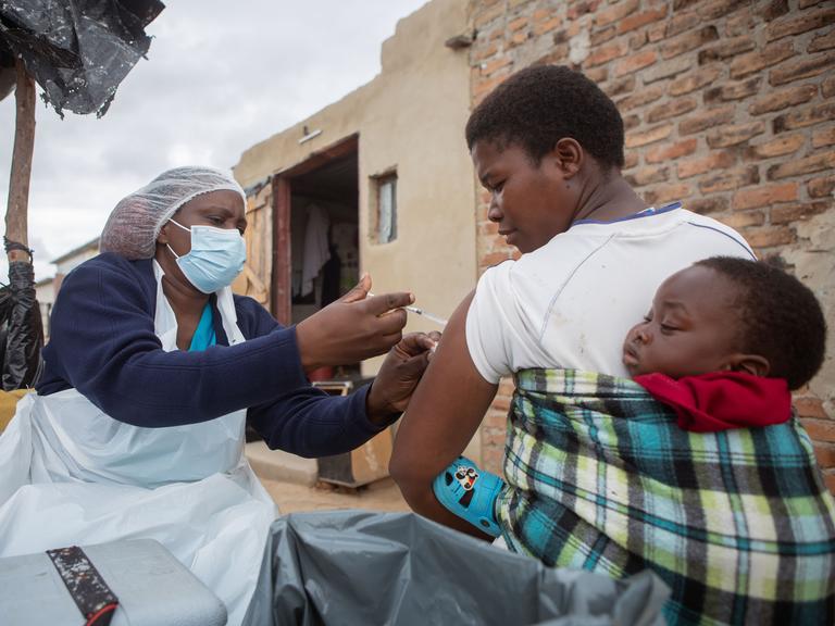 Eine Krankenschwester impft in Zimbabwe eine Mutter, die ihr Kind auf dem Rücken trägt.  