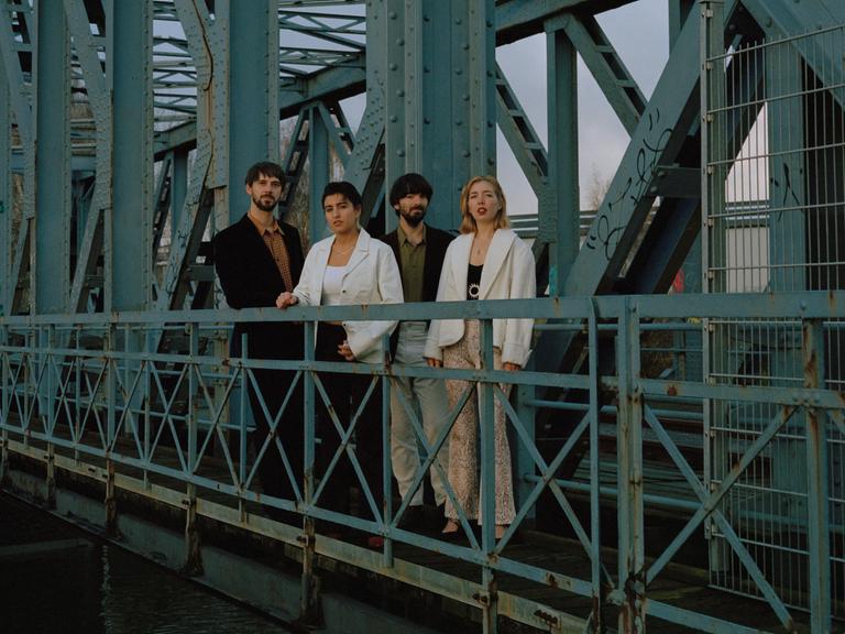 Vier Personen stehen auf einer alten, rostigen Brücke und schauen über die Brüstung in die Kamera.