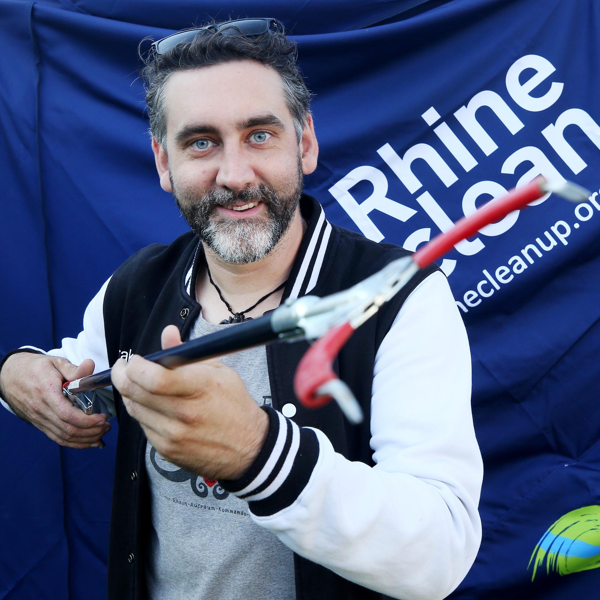 Christian Stock steht vor einem Transparent der Aktion "Rhine Cleanup" und zeigt einen Müllsammelstock.