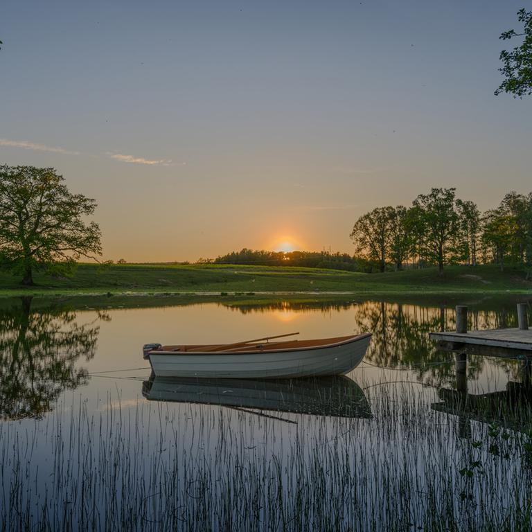 Zeitebenen und Perspektiven wandeln sich, Identitäten verschwimmen in einer schwedischen Sommernacht.. Zu sehen: Auf einem See ist ein Boot neben einem Steg. Eine Wiese mit Bäumen ist im Hintergrund  und ein Sonnenuntergang ist zu sehen. 