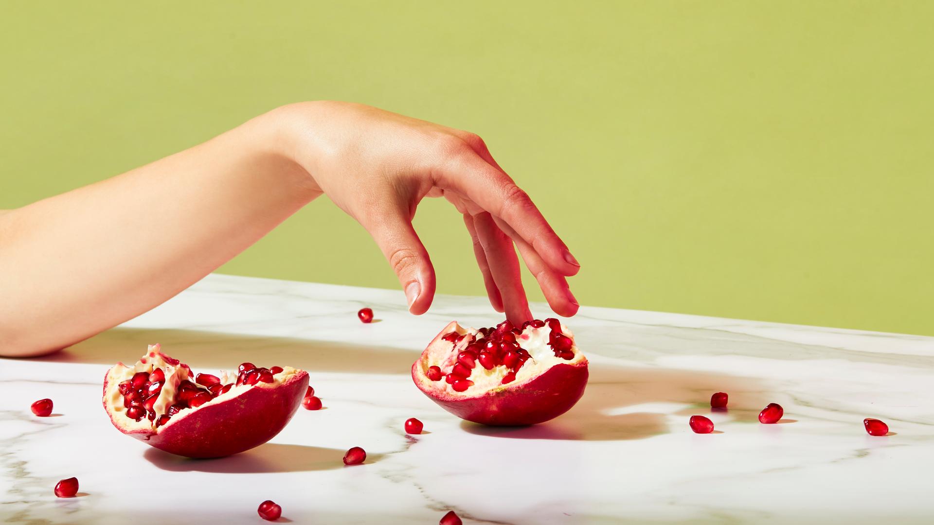 Ein geöffneter Granatapfel liegt auf einem Tisch. Einzelne Kerne sind verstreut. Eine Hand berührt sanft einen Teil des Granatapfels. Das Bild mutet sinnlich an. Ein geöffneter Granatapfel liegt auf einem Tisch. Einzelne Kerne sind verstreut. Eine Hand berührt sanft einen Teil des Granatapfels. Das Bild mutet sinnlich an.