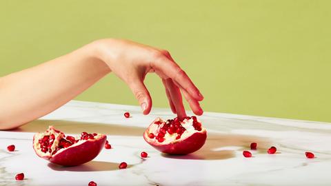 Ein geöffneter Granatapfel liegt auf einem Tisch. Einzelne Kerne sind verstreut. Eine Hand berührt sanft einen Teil des Granatapfels. Das Bild mutet sinnlich an.