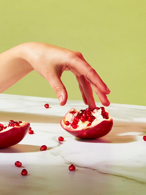 Ein geöffneter Granatapfel liegt auf einem Tisch. Einzelne Kerne sind verstreut. Eine Hand berührt sanft einen Teil des Granatapfels. Das Bild mutet sinnlich an.