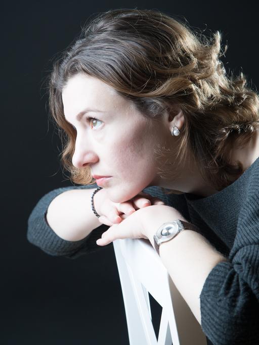 Tetiana Goncharuk sitzt verkehrt herum auf einem Stuhl und stützt ihren Kopf auf die Stuhllehne. Der Blick geht in die Ferne. Sie trägt einen dunkelgrauen Pullover und Perlohringe.