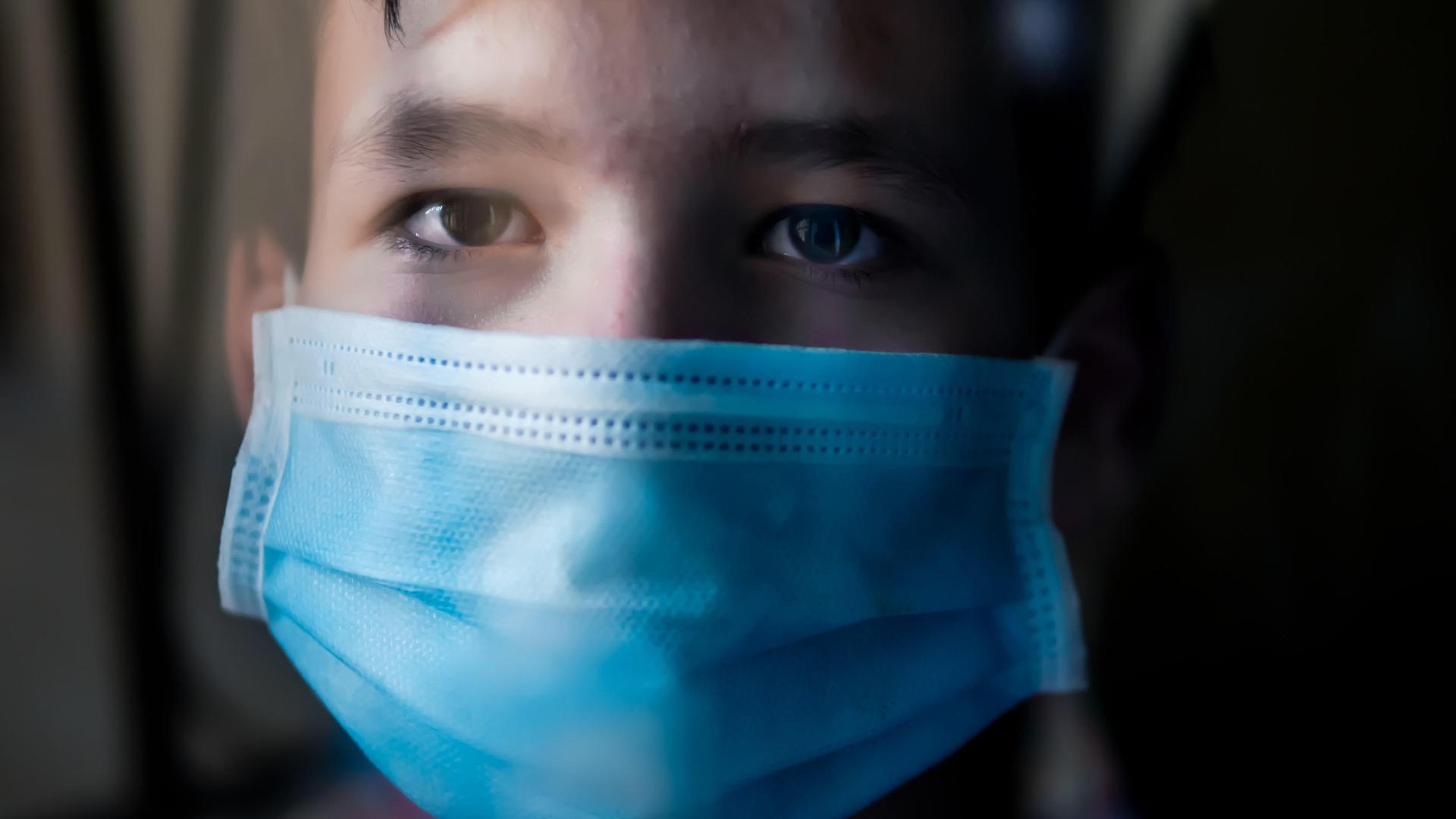 Ein Junge mit medizinischer Gesichtsmaske und traurigem Blick hinter einem Fenster. Ein Junge mit medizinischer Gesichtsmaske und traurigem Blick hinter einem Fenster.
