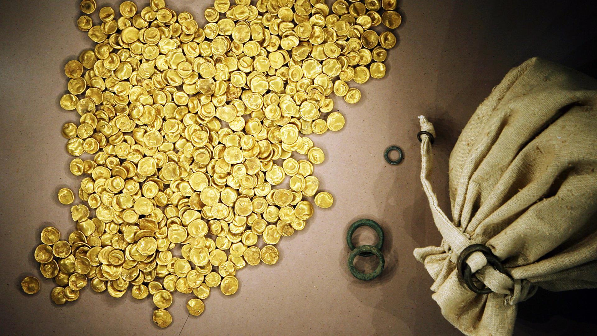 Viele Gold-Münzen liegen neben einem offenen Stoff-Beutel.