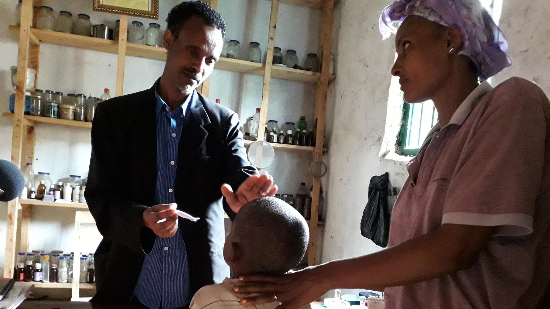 Der Heiler Tegistu behandelt in Äthiopien einen fünfjährigen Jungen, der an einer Pilzerkrankung leidet. Er hat seine Hand auf dem Kopf des Jungen. Daneben steht dessen Mutter. Im Hintergrund sind Gläser mit Pflanzen.