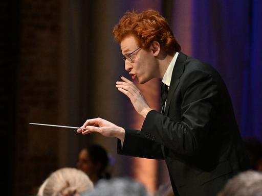 Ein junger Mann mit Brille und roten kurzen Haaren gibt ein Zeichen, dass das Orchester leiser spielen soll. In der rechten Hand hält er einen Dirigierstab.