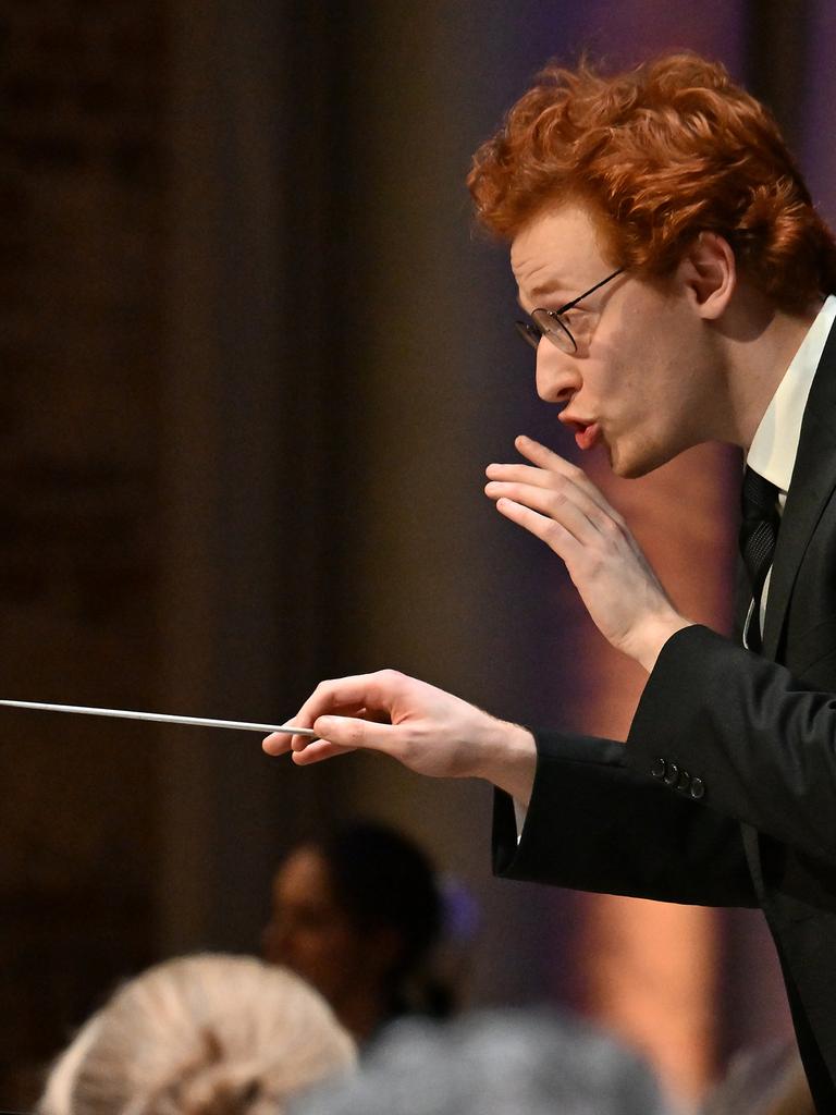 Ein junger Mann mit Brille und roten kurzen Haaren gibt ein Zeichen, dass das Orchester leiser spielen soll. In der rechten Hand hält er einen Dirigierstab.