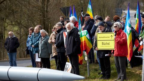 Zum Auftakt einer Friedensaktion im März 2018 am Fliegerhorst Büchel haben sich Demonstranten an dem Fliegerhosrt versammelt. Im Vordergrund ein schematisch nachgebaute Rakete. Im Hintergrund stehen Menschen, zum Teil halten sie Friedensymbole wie die Regenbogenfahne.
