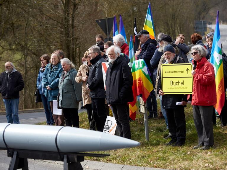 Zum Auftakt einer Friedensaktion im März 2018 am Fliegerhorst Büchel haben sich Demonstranten an dem Fliegerhosrt versammelt. Im Vordergrund ein schematisch nachgebaute Rakete. Im Hintergrund stehen Menschen, zum Teil halten sie Friedensymbole wie die Regenbogenfahne.