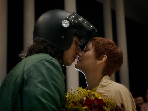 Ein Mann mit Motorradhelm küsst eine Frau, die einen Blumenstrauß hält.