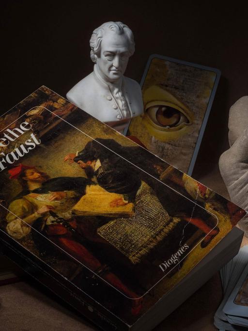 Hörspielkomödie über Goethes "Faust" und die Mühen des Deutschwerdens. Zu sehen: Stilleben mit Büchern, Glaskugel, Tarotkarten, Goethe-Büste, das Buch "Faust" und einer Plastikhand zu einer Faust geballt. 