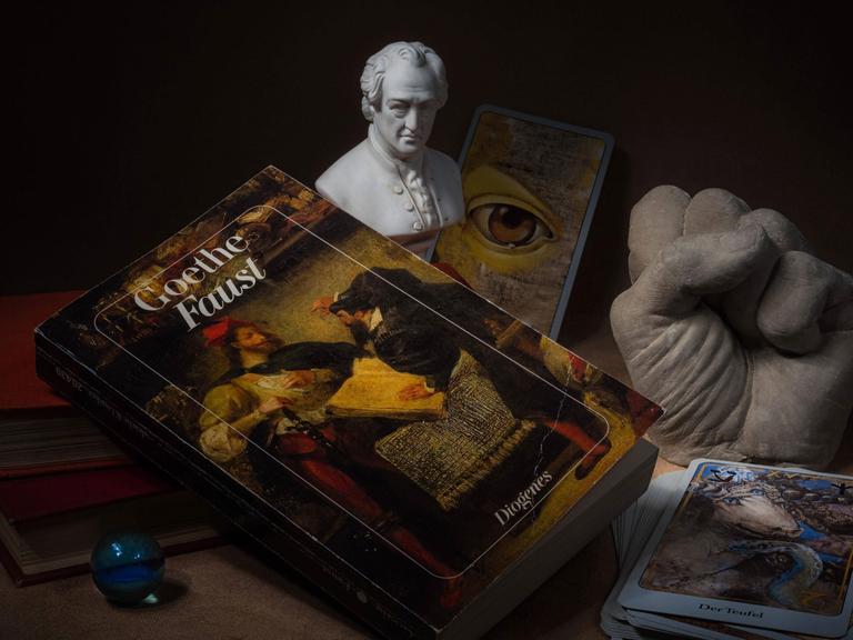 Hörspielkomödie über Goethes "Faust" und die Mühen des Deutschwerdens. Zu sehen: Stilleben mit Büchern, Glaskugel, Tarotkarten, Goethe-Büste, das Buch "Faust" und einer Plastikhand zu einer Faust geballt. 