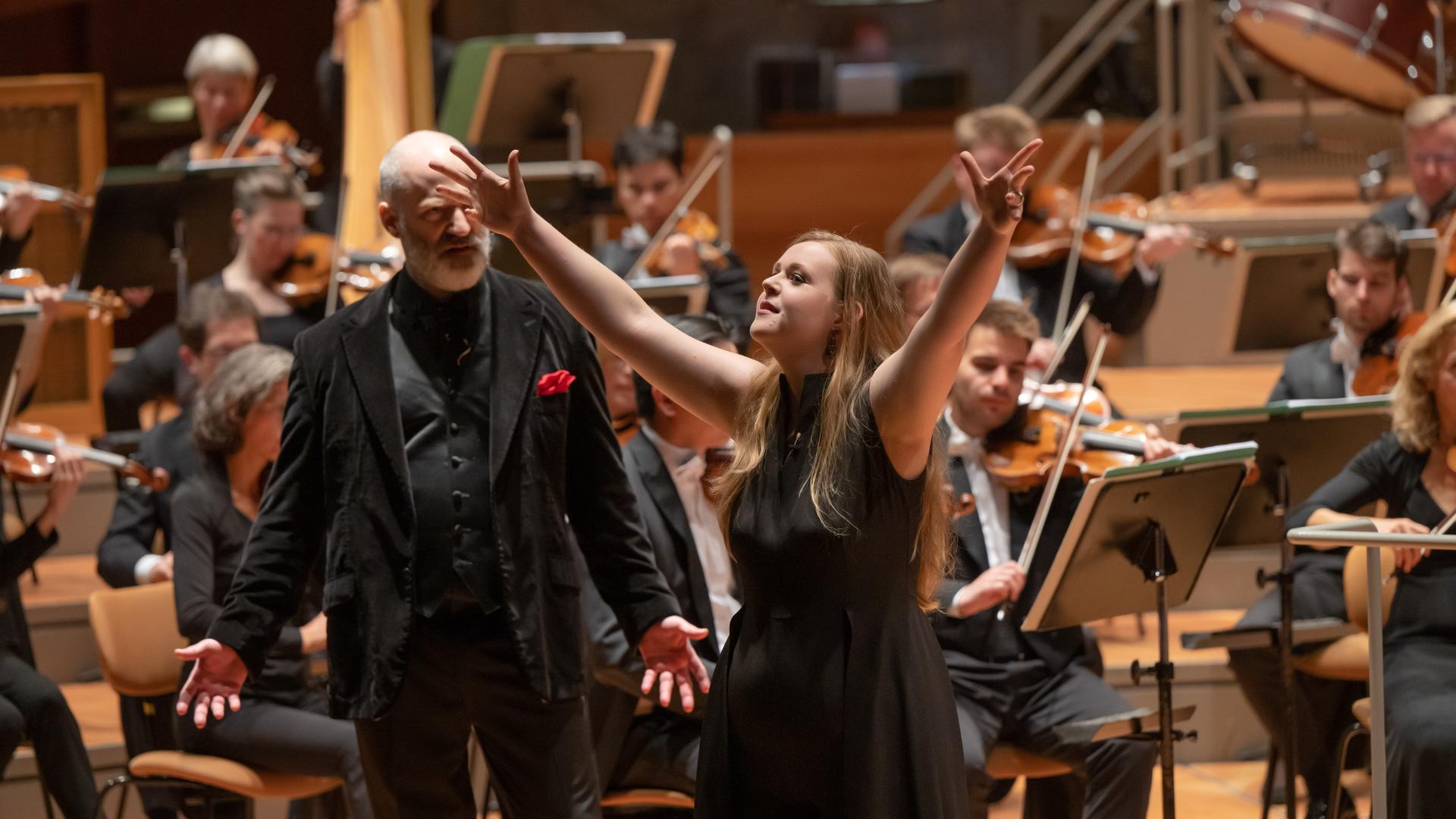 Eine Frau hebt beide Arme hin die Höhe, sie blickt triumphierend. Hinter steht ein älterer Mann, der ernst blickt. Beide stehen vor einem großen Symphonieorchester. Eine Frau hebt beide Arme hin die Höhe, sie blickt triumphierend. Hinter steht ein älterer Mann, der ernst blickt. Beide stehen vor einem großen Symphonieorchester.