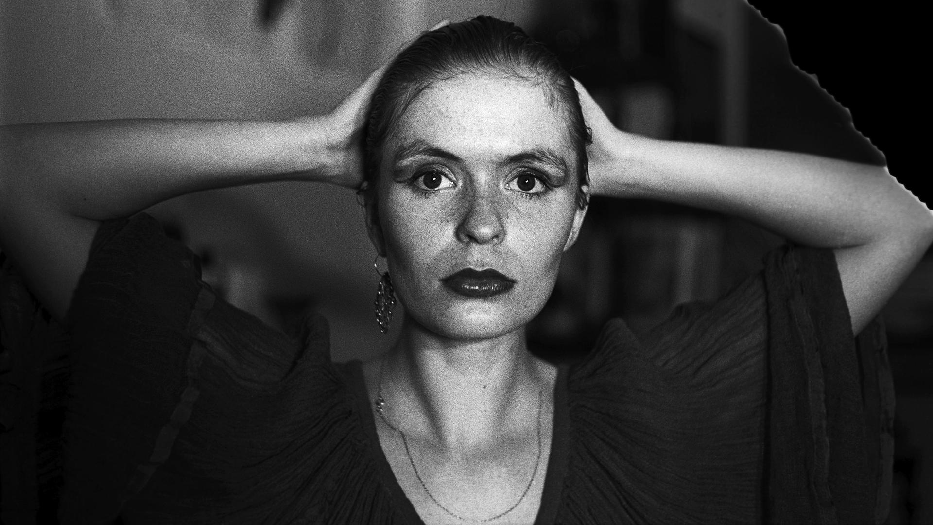 Aus der Serie "Lange Weile, 1983-1989" von der Fotografin Tina Bara. Eine Frau faltet die Hände hinter ihrem Kopf zusammen und blickt direkt in die Kamera. Aus der Serie "Lange Weile, 1983-1989" von der Fotografin Tina Bara. Eine Frau faltet die Hände hinter ihrem Kopf zusammen und blickt direkt in die Kamera.