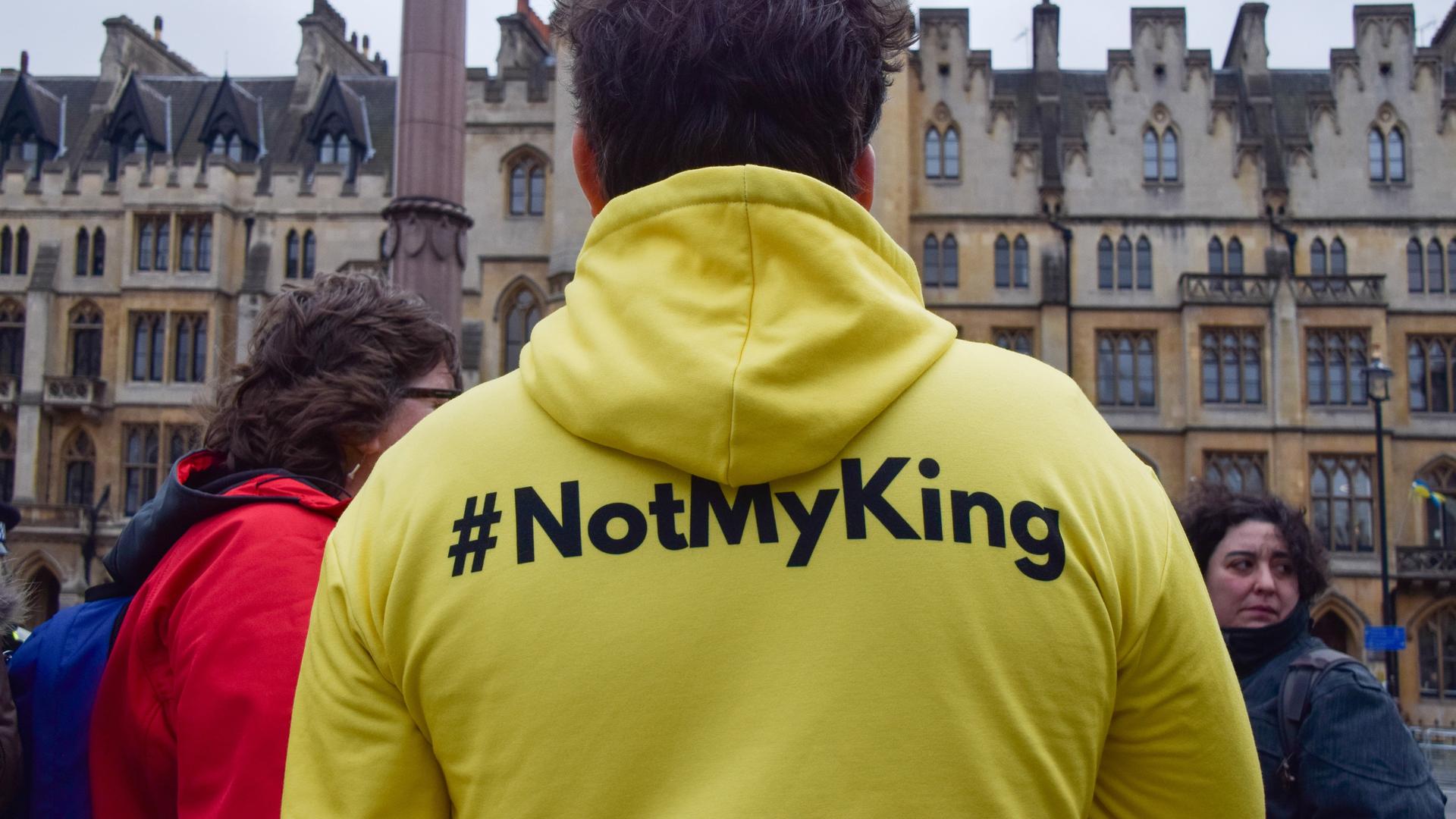 Eine Person, die einen neongelben Pullover mit der Aufschrift "#notmyking" trägt steht zusammen mit anderen Personenn vor einem Gebäude in London Eine Person, die einen neongelben Pullover mit der Aufschrift "#notmyking" trägt steht zusammen mit anderen Personenn vor einem Gebäude in London