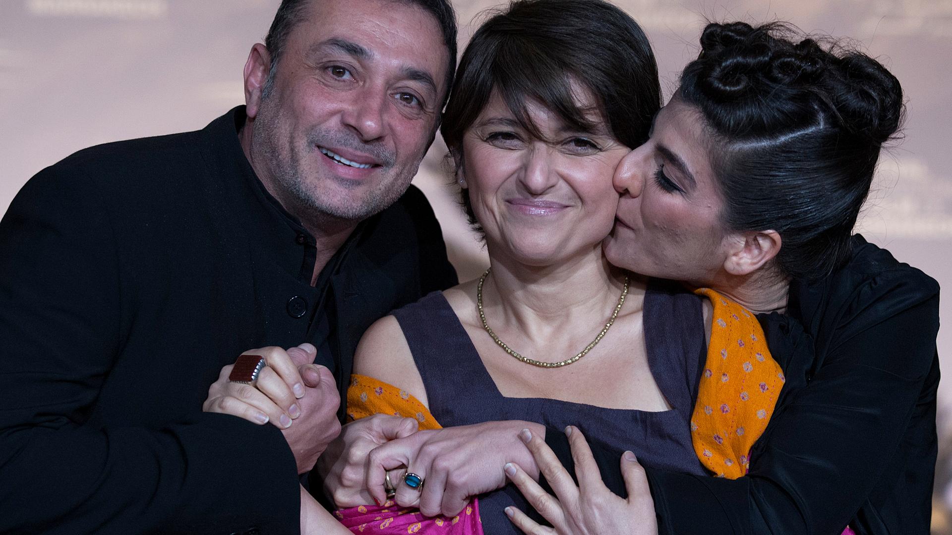 Sepideh Farsi steht zwischen einem Mann und einer Frau, die sie umarmen. Sie selbst lächelt in die Kamera. Sepideh Farsi steht zwischen einem Mann und einer Frau, die sie umarmen. Sie selbst lächelt in die Kamera.