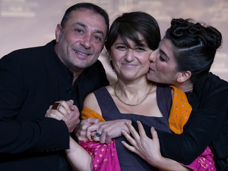 Sepideh Farsi steht zwischen einem Mann und einer Frau, die sie umarmen. Sie selbst lächelt in die Kamera.