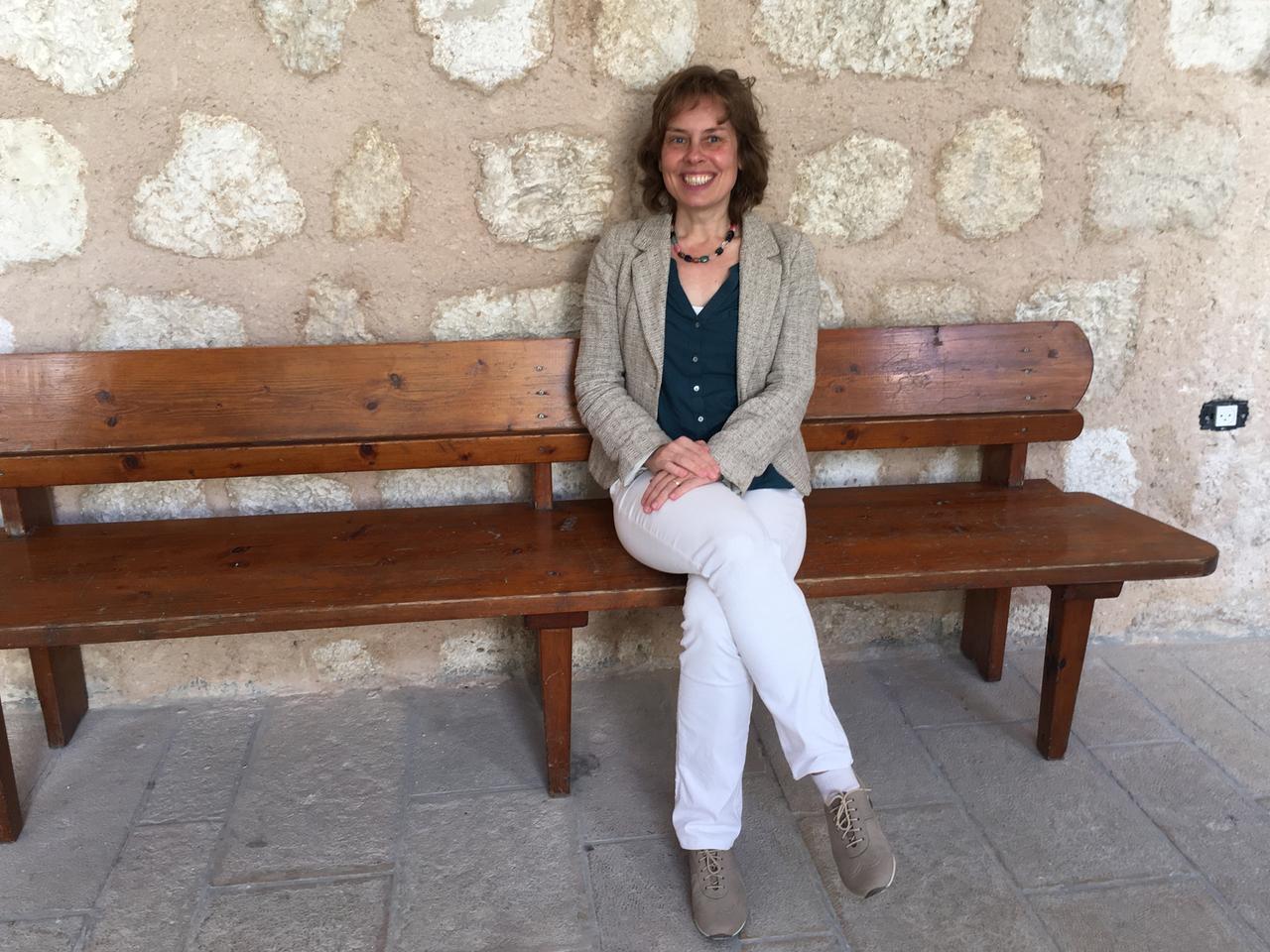 Eine braunhaarige Frau in blauer Blouse und mit grauem Blazer sitzt mit überschlagenen Beinen auf einer Holzbank vor einer Steinmauer und lächelt strahlend in die Kamera. Eine braunhaarige Frau in blauer Blouse und mit grauem Blazer sitzt mit überschlagenen Beinen auf einer Holzbank vor einer Steinmauer und lächelt strahlend in die Kamera.