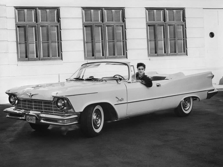 Der Sänger Elvis Presley in seinem Chrysler Imperial auf dem Studiogelände der Filmproduktionsfirma MGM in Hollywood, USA (1958). Sein Auto ist ein großes, weißes Cabriolet, das Verdeck ist geöffnet.
