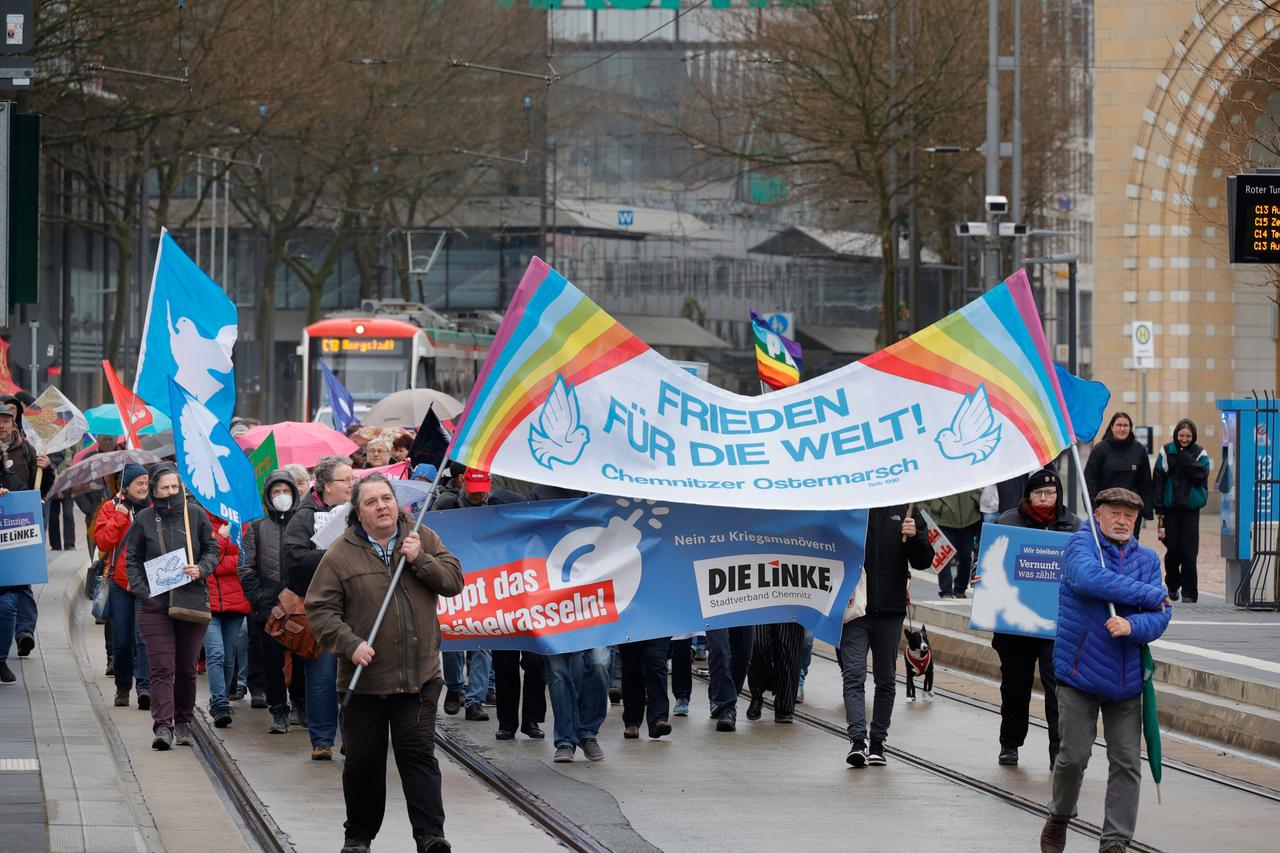 Dutzende Teilnehmer gehen über eine Straße. Sie tragen Transparente. Hinter ihnen fährt eine Straßenbahn. Das Wetter ist eher trübe. Dutzende Teilnehmer gehen über eine Straße. Sie tragen Transparente. Hinter ihnen fährt eine Straßenbahn. Das Wetter ist eher trübe.