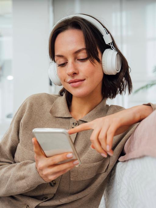 Eine junge Frau sitzt mit Kopfhörern lässig auf einer hellen Couch und tippt lächelnd auf ihrem Handy. 
