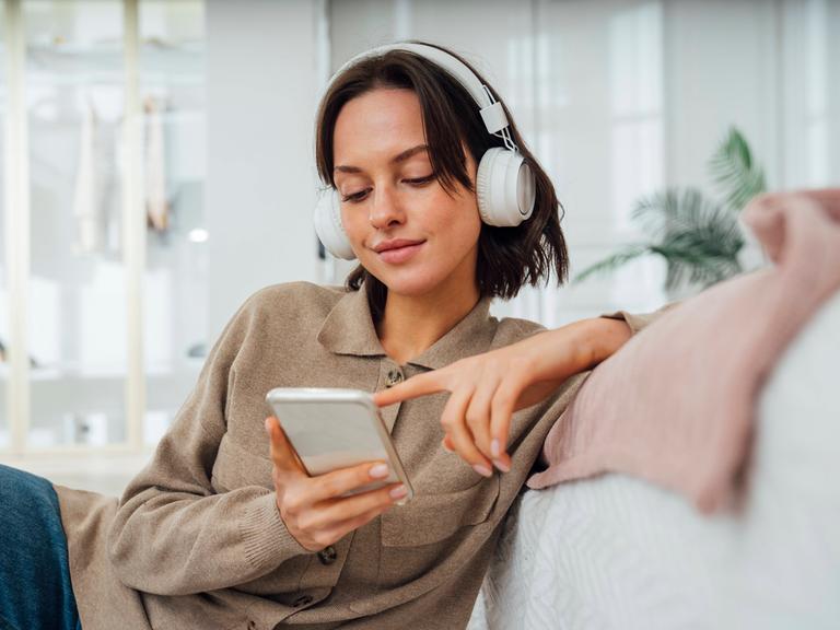 Eine junge Frau sitzt mit Kopfhörern lässig auf einer hellen Couch und tippt lächelnd auf ihrem Handy. 
