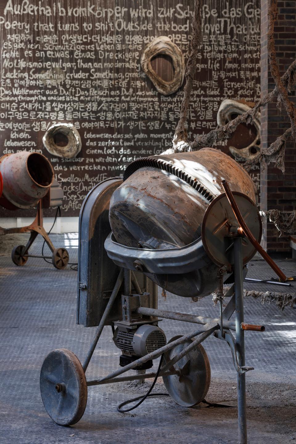 Blick in die Ausstellung: Zwei Betonmischer vor einem Wandfedicht auf Deutsch, Englisch und Koreanisch. Blick in die Ausstellung: Zwei Betonmischer vor einem Wandfedicht auf Deutsch, Englisch und Koreanisch.