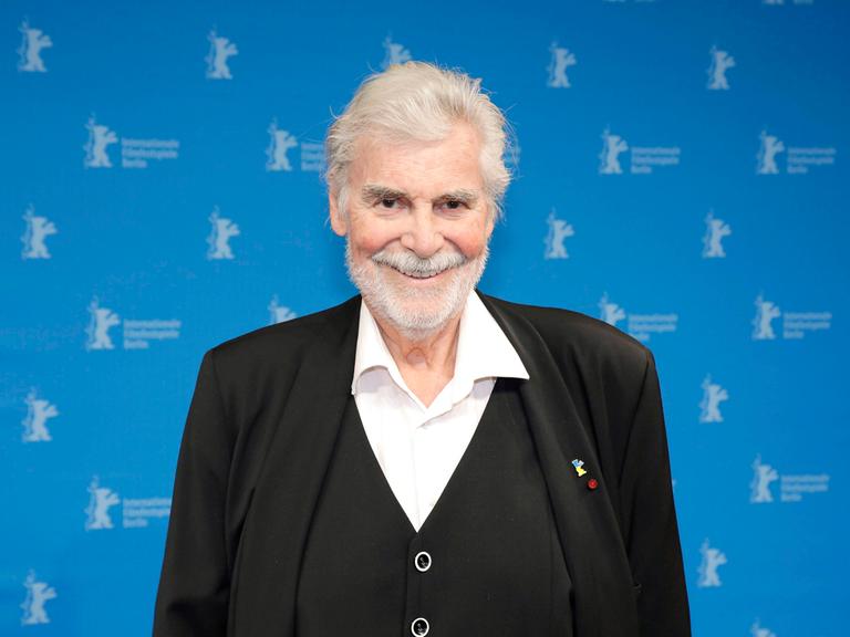 Peter Simonischek steht im schwarzen Anzug vor einem blauen Hintergrund mit kleinen Berlinale Logos.