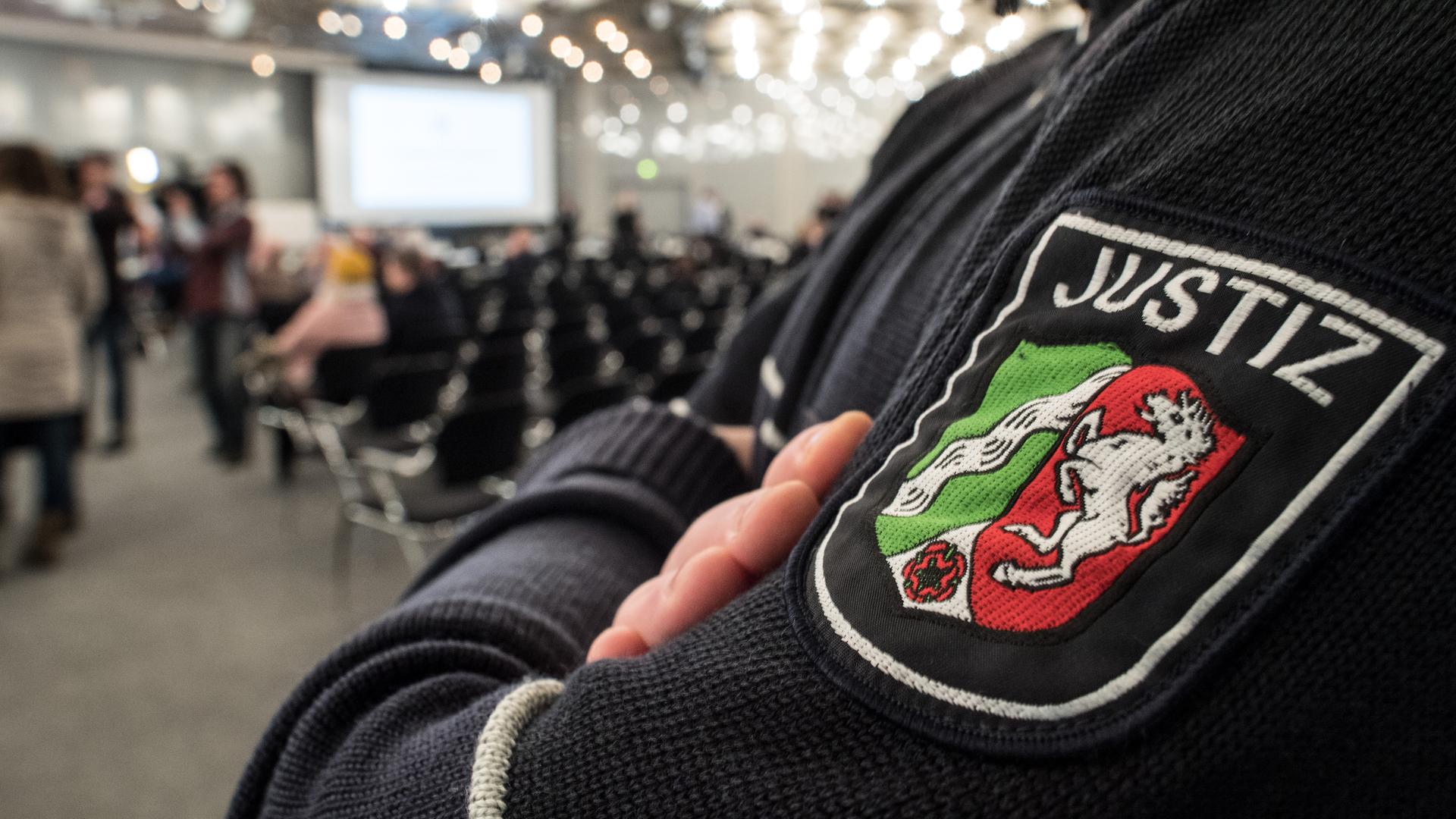 Blick auf das Justiz-Symbol auf dem Ärmel eines Justiz-Mitarbeiters in Düsseldorf. Im Hintergrund: Blick in den Saal. Blick auf das Justiz-Symbol auf dem Ärmel eines Justiz-Mitarbeiters in Düsseldorf. Im Hintergrund: Blick in den Saal.