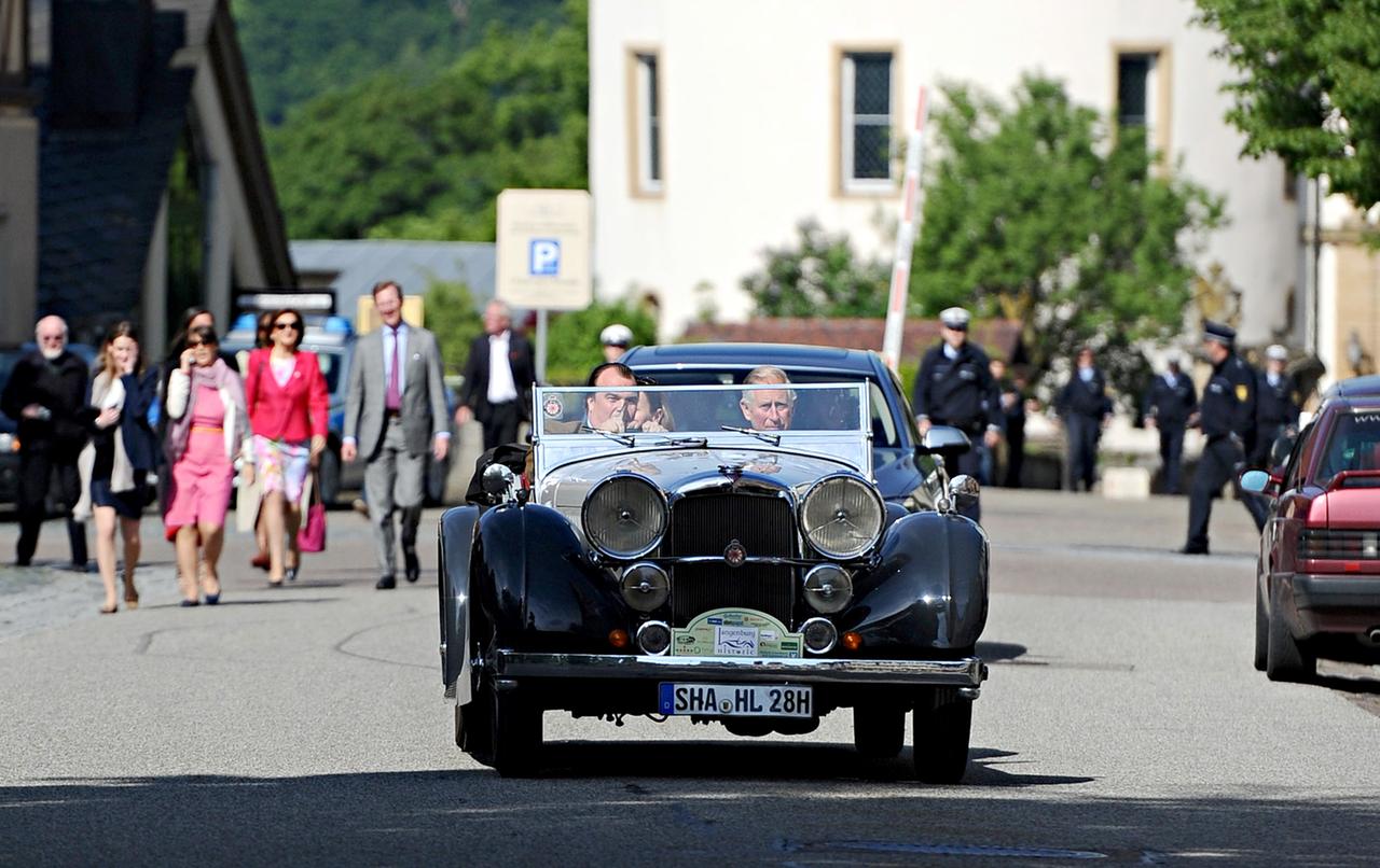 Der britische Thronfolger Prinz Charles sitzt mit einem Mann in Oldtimer-Cabrio. Sie fahren durch einen kleinen Ort.