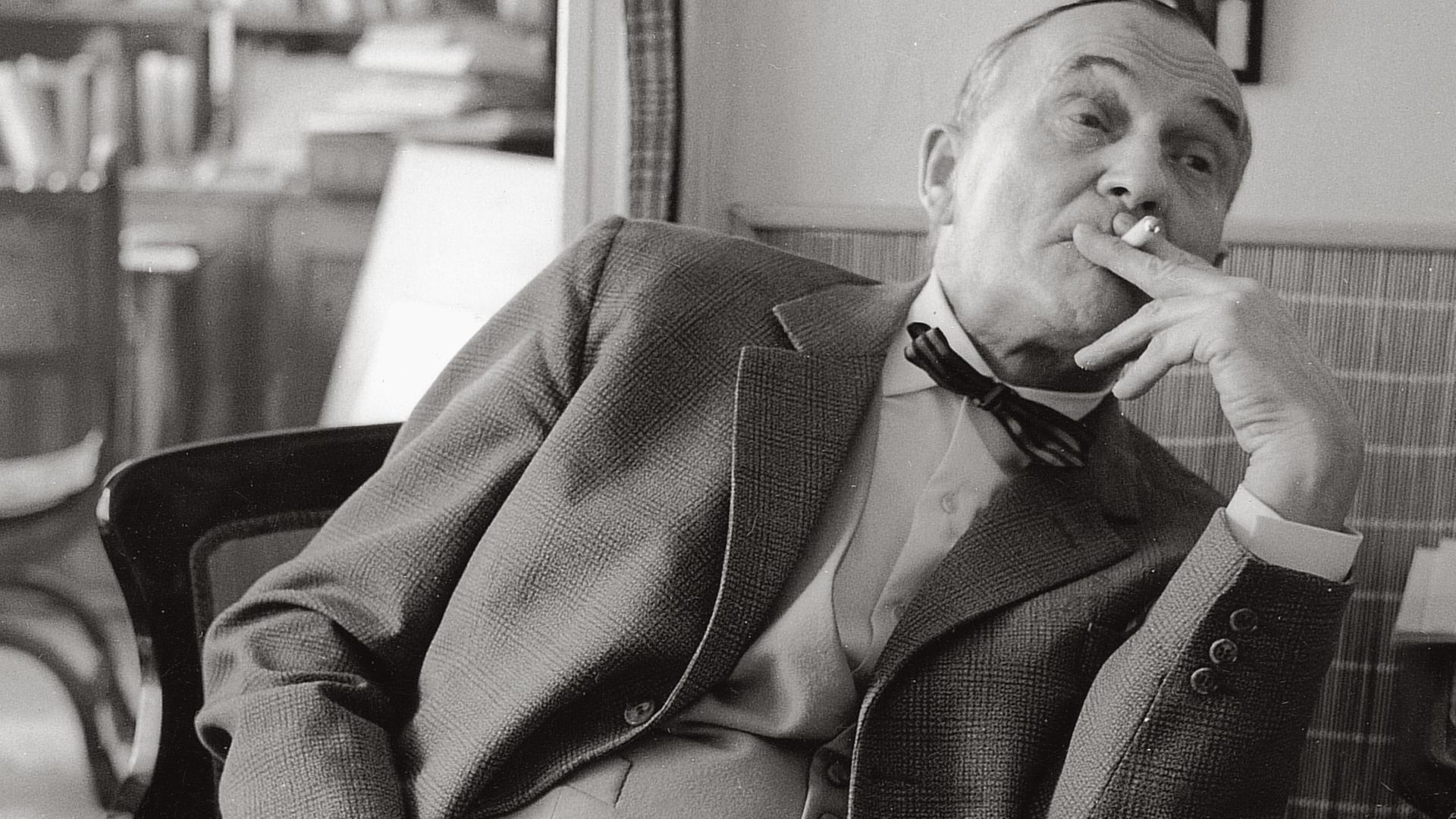 Heimito von Doderer zurückgelehnt auf einem Stuhl sitzend und Zigarette rauchend in seinem Wohnhaus in Wien.