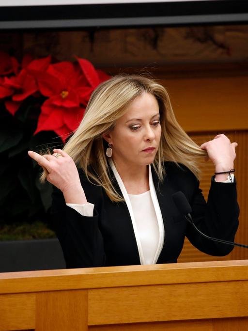 Giorgia Meloni steht an einem Pult im italienischen Parlament und fährt sich mit den Händen durch die langen blonden Haare.