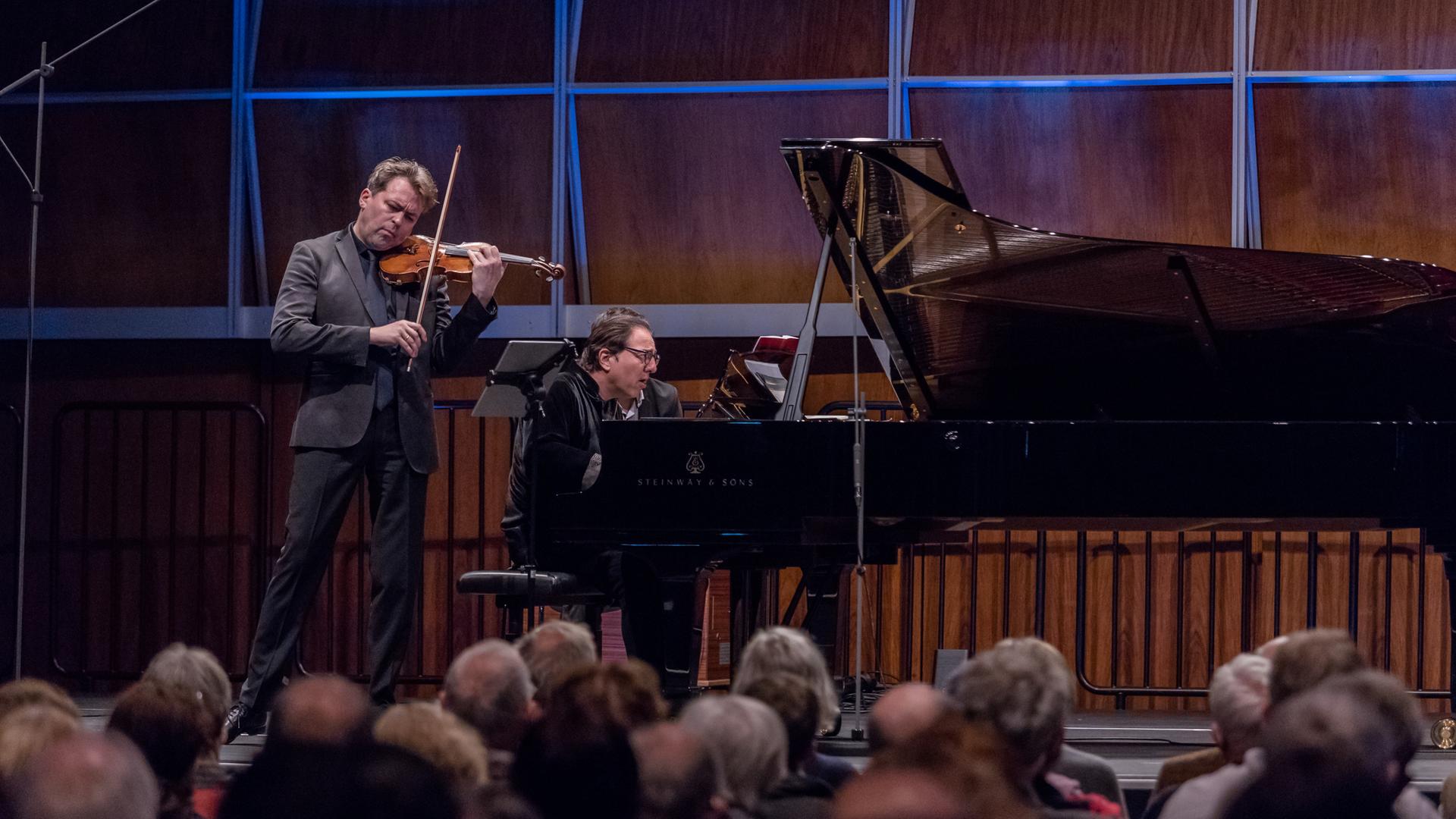 Ein Geiger und ein Pianist geben ein Konzert in einem voll besetzten, holzvertäfelten Saal, der blau beleuchtet ist. Ein Geiger und ein Pianist geben ein Konzert in einem voll besetzten, holzvertäfelten Saal, der blau beleuchtet ist.