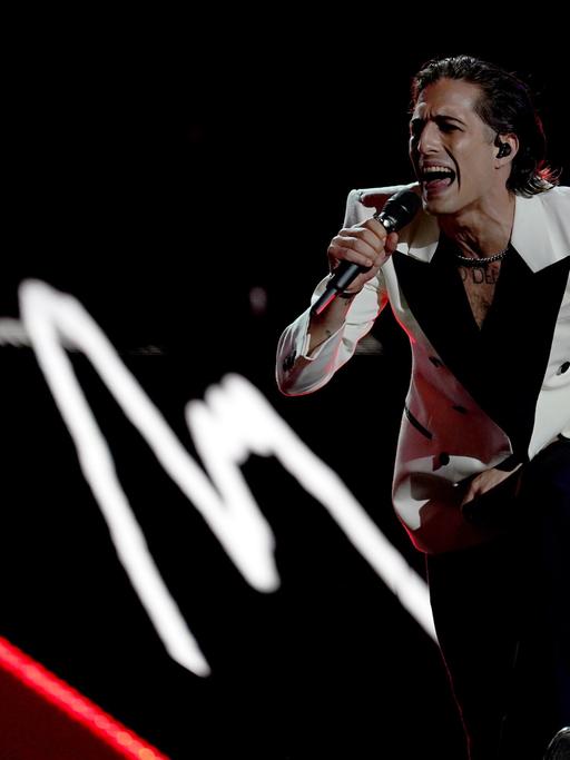 Måneskin-Sängder Damiano David posiert auf der Bühne. Er trägt ein weißes Jacket mit schwarzem Kragen und eine dunkle Hose und singt, weit vorgebeugt, kraftvoll ins Mikrofon.