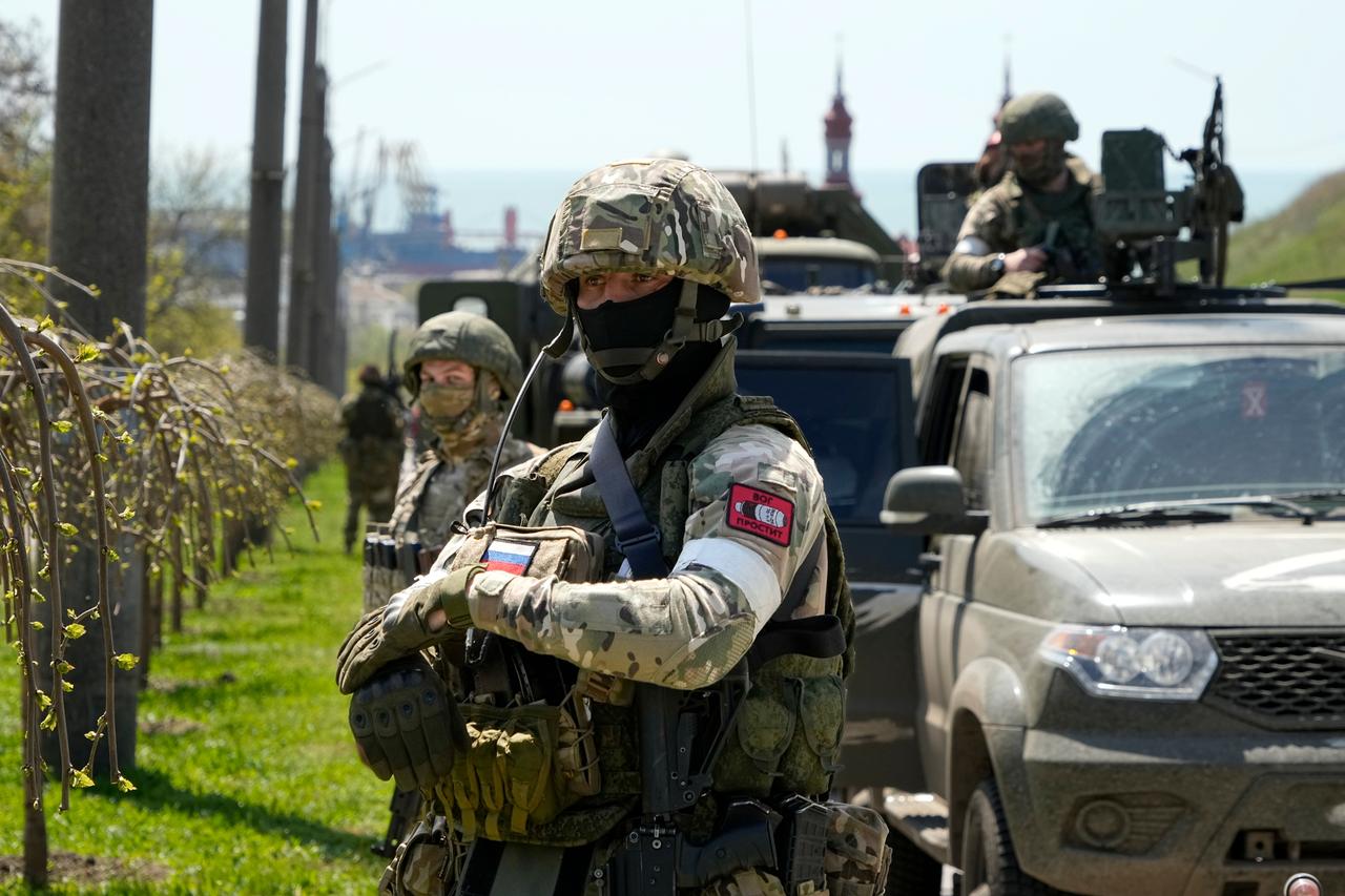 Zwei Soldaten in Kampfmonitur und mit Masken stehen an einer Straße neben einem militärischen Fahrzeugkonvoi. 