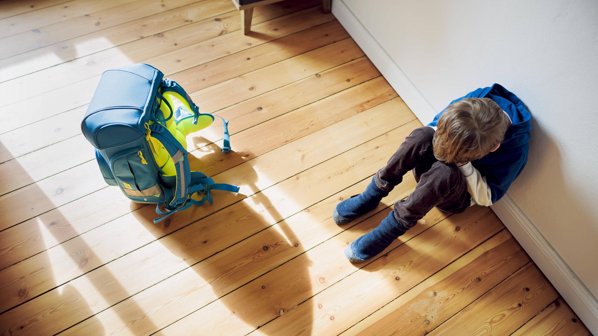  Ein Junge sitzt traurig in seinem Kinderzimmer neben seinem Schulranzen und versteckt sein Gesicht hinter seinen Händen.