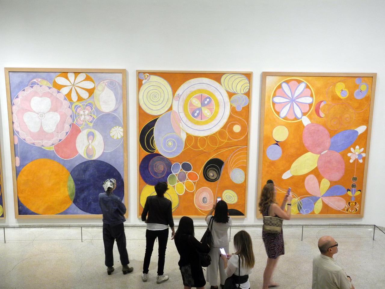In einem Museum schauen sich Besucher drei große Gemälde der schwedischen Malerin Hilma af Klint an. Es sind abstrakte Bilder. In einem Museum schauen sich Besucher drei große Gemälde der schwedischen Malerin Hilma af Klint an. Es sind abstrakte Bilder.