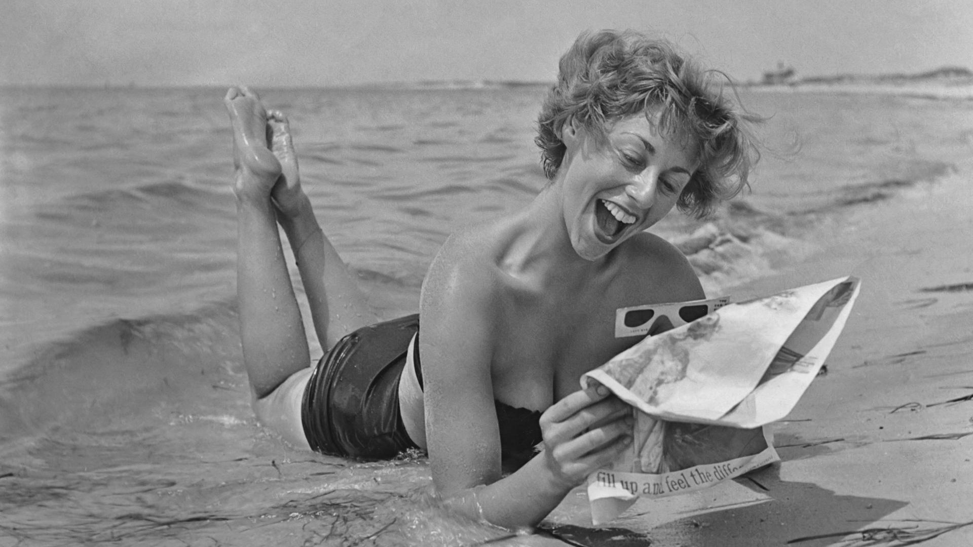 Schwarzweißfoto einer Frau, die im Bikini am Strand liegt und eine Zeitung liest. Schwarzweißfoto einer Frau, die im Bikini am Strand liegt und eine Zeitung liest.
