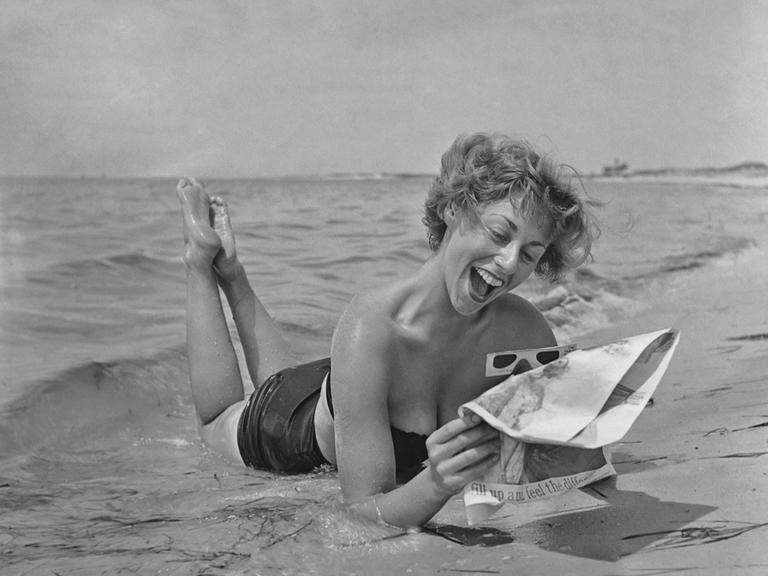 Schwarzweißfoto einer Frau, die im Bikini am Strand liegt und eine Zeitung liest.