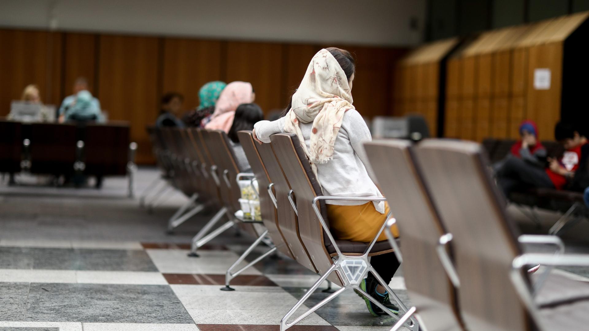 Ein paar Frauen sitzen im Wartebereich einer Behörde. Sie sind von hinten zu sehen und tragen Kopftücher.