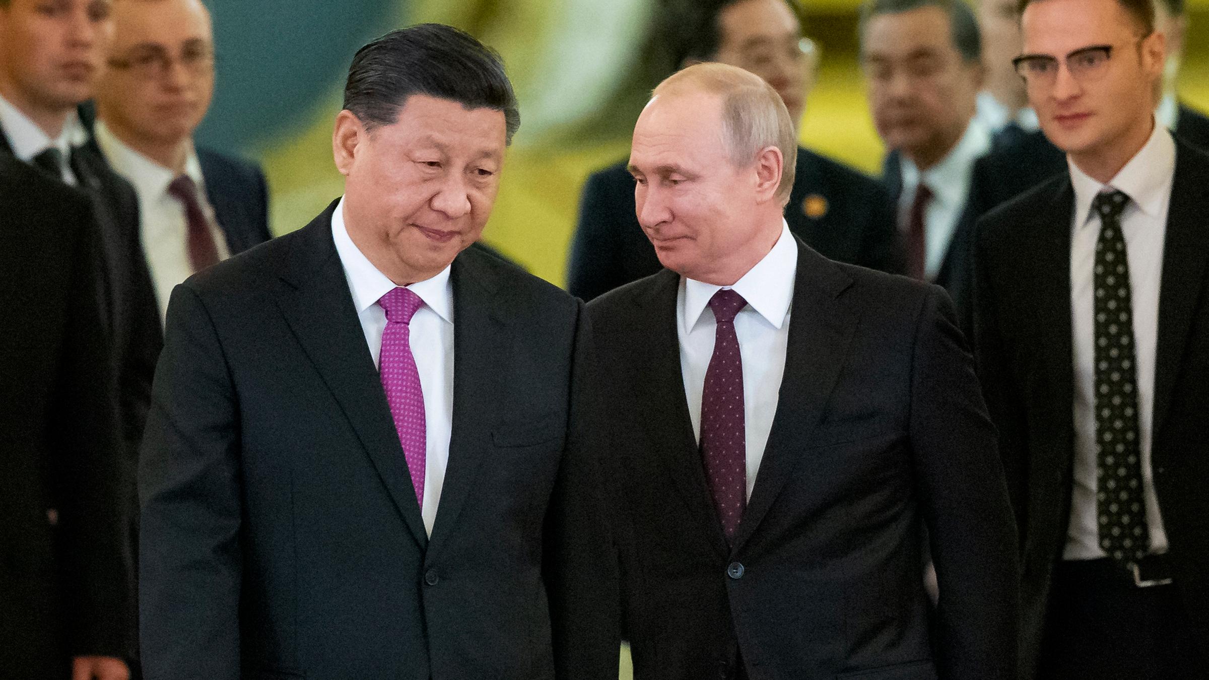 Chinas Präsident Xi Jinping und Russlands Staatschef Wladimir Putin gehen in schwarzen Anzügen gekleidet nebeneinander her, hinter ihnen weitere Männer in schwarzen Anzügen.